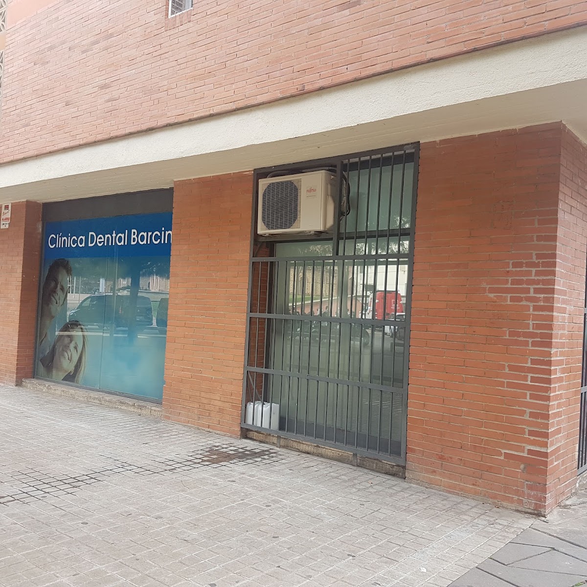 Fachada de la clínica: ventanal con vinilo azul rotulado Clínica Dental Barcin, con la última letra tapada por el pilar de ladrillo, sobre pared de ladrillo rojo; a la derecha, una ventana enrejada con un equipo de aire acondicionado encima.